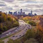 Photo of a highway along fall-colored trees and a city with skyscrapers in the distance.