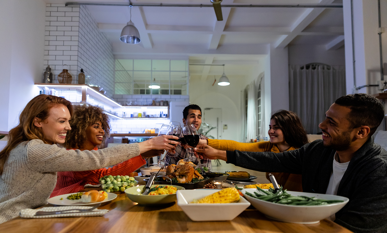 Side view of a group of young adult multi-ethnic male and female friends sitting at a table at home set for Thanksgiving dinner making a toast with glasses of red wine.