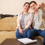 Positive family couple shows keys bunch near coffee table with apartment rental contract sitting with cute cat on sofa in light room.