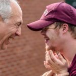 An elderly white man with gray hair and a gray colored shirt laughs with a younger male with strawberry blond hair wearing a maroon baseball cap and matching shirt.