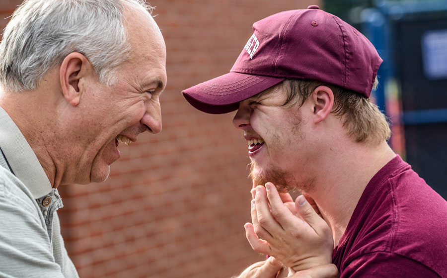 An elderly white man with gray hair and a gray colored shirt laughs with a younger male with strawberry blond hair wearing a maroon baseball cap and matching shirt.