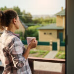A white female holding a pink coffee cup and wearing a pink-gray plaid shirt looks at a couple yellow houses from a second floor balcony. She has her curly hair done up in a ponytail.
