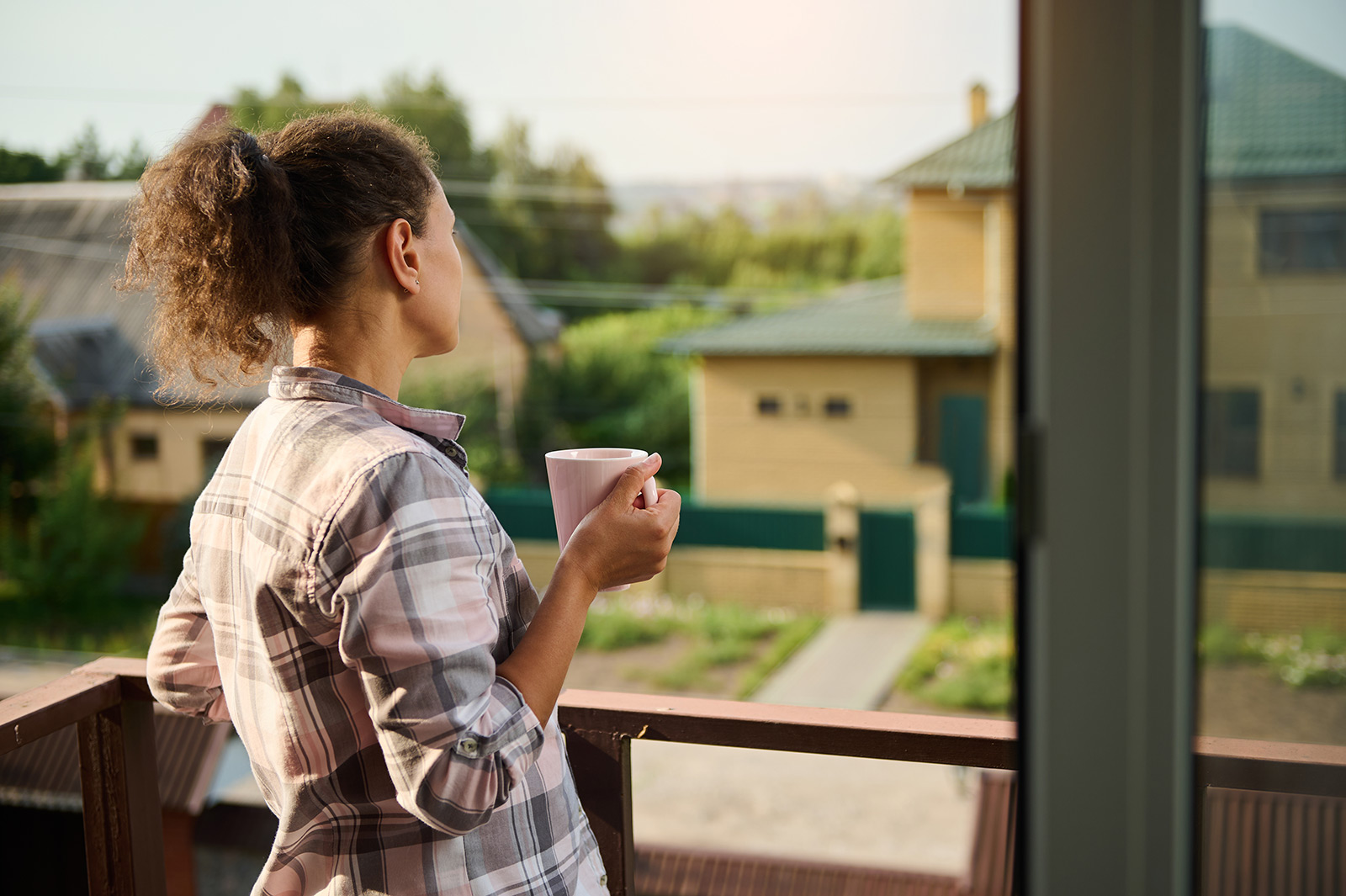 A white female holding a pink coffee cup and wearing a pink-gray plaid shirt looks at a couple yellow houses from a second floor balcony. She has her curly hair done up in a ponytail.