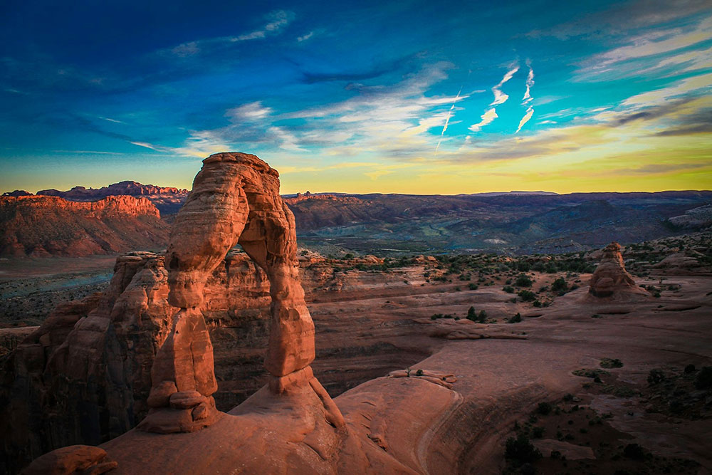 Delicate Arch at Arches National Park in Utah, glowing in the evening light with red rock formations and a colorful desert sky in the background.