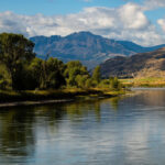 Scenic view of South Central Montana featuring a calm river, lush trees, and distant mountains under a partly cloudy sky.