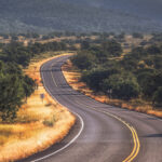 Winding road through juniper-dotted grasslands with forested hills in the distance near Payson, Central Arizona, on a clear day.